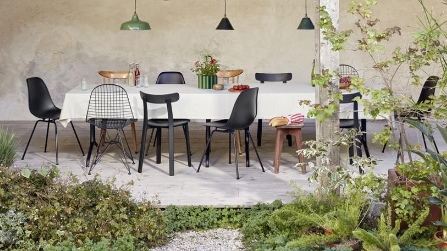 Auf dem Bild sind im Vordergrund links und rechts Pflanzen zu sehen. In der Mitte steht ein Tisch auf einer Terrasse. Um den Tisch herum stehen verschiedene Stühle der Marke Vitra.